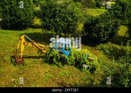 Un vecchio trattore con una benna, abbandonato da un giardino verde Foto Stock