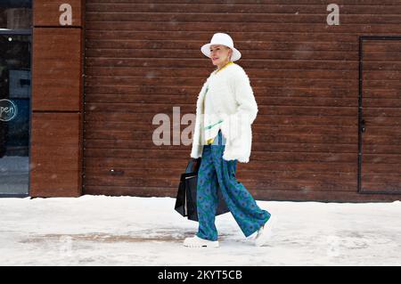 Donna matura alla moda che cammina sullo sfondo del muro in strada invernale con borse per la spesa, capelli corti, vestendo il cappello fedora ed elegante Foto Stock