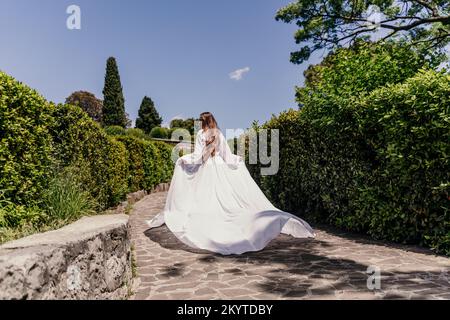 una bella donna con lunghi capelli castani e lungo vestito bianco corre lungo un sentiero lungo splendidi cespugli nella vista posteriore del parco Foto Stock