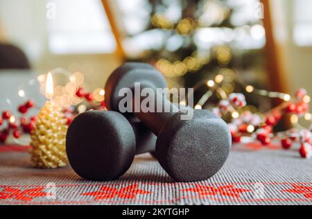 Stile di vita sano durante il periodo di festa di Natale concetto. Manubri neri con ornamenti natalizi albero di Natale sfocato con le luci di Natale accese. Foto Stock