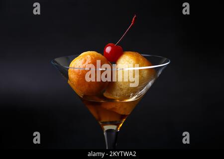 Rum baba napoletano (o baba au rum) in un bicchiere di martini con una ciliegina da cocktail su sfondo nero. Piccole torte di lievito imbevute di sciroppo di rum. Tradizione Foto Stock