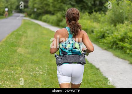 Foto sul retro scattata da una bruna vestita di abbigliamento sportivo che pratica da sola una sessione cardio facendo jogging all'aperto nel parco Foto Stock