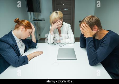 Donna bionda, dai capelli rossi e uomo bearded in vestiti nell'ufficio.i colleghi siedono ad un tavolo in una sala di conferenza e tengono le loro teste nel pensiero. Foto Stock