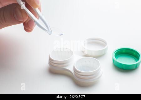 Mani che raccolgono una lente a contatto con pinzette e materiale di manutenzione per lenti a contatto su tavolo bianco. Salute e cura degli occhi, vista e visione, op Foto Stock