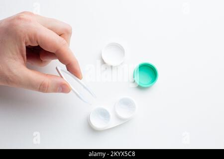 Mani che raccolgono una lente a contatto con pinzette e materiale di manutenzione per lenti a contatto su tavolo bianco. Salute e cura degli occhi, vista e visione, op Foto Stock