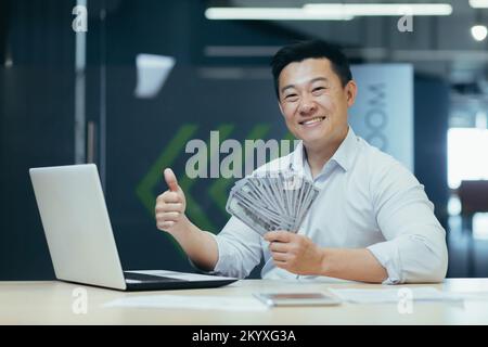 Ritratto di un giovane uomo d'affari asiatico bello, capo, SEO milionario. Si siede in ufficio al tavolo con un computer portatile, tiene denaro in contanti nelle mani, sorride alla fotocamera, punta un super dito. Foto Stock