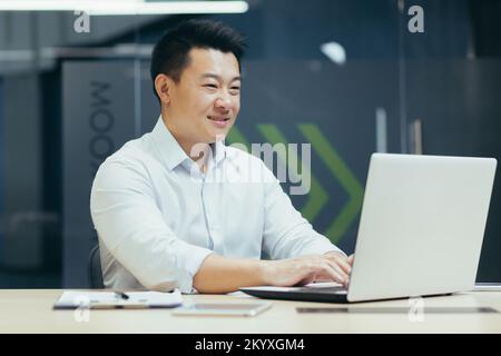 Ritratto di un giovane uomo d'affari asiatico sorridente che lavora in ufficio, seduto a un tavolo, a digitare su un computer portatile. Foto Stock