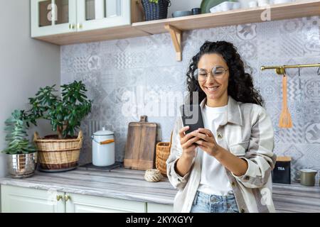 Giovane donna ispanica in piedi a casa in cucina con telefono. Effettua acquisti online, ordini, consegne a domicilio. Foto Stock