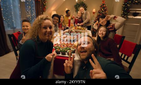 Donna caucasica con bastone selfie scatta foto di gruppo di famiglia. Famiglia multiculturale che celebra Natale o Capodanno 2023. Tavolo con piatti. Atmosfera di famiglia cena di Natale a casa. Vista telecamera. Foto Stock