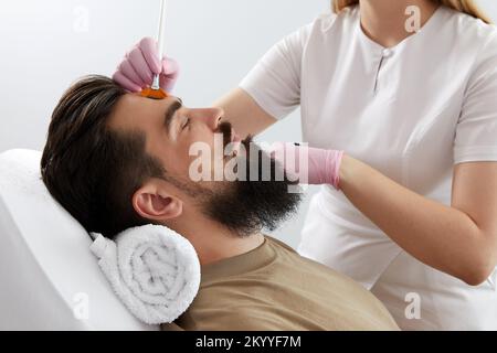 Cosmetologo applicare una maschera di argilla al viso maschile con pennello. Trattamento termale e cura del viso nel salone di bellezza Foto Stock