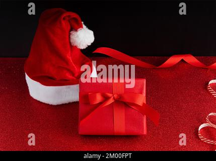 Sfondo di Natale con un cappello di babbo natale e una scatola regalo rossa con nastro su una superficie di glitter e uno sfondo scuro. Foto Stock