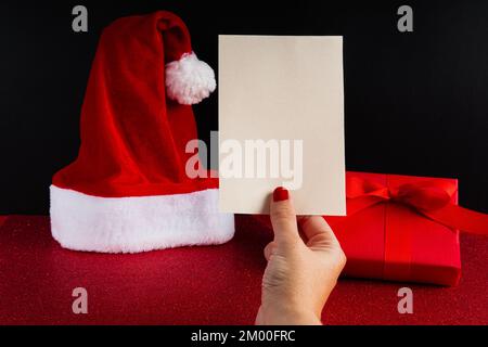 Mano della donna con le unghie rosse che tengono una cartolina vuota sopra un insieme di natale con un cappello di babbo natale ed una scatola rossa completa del regalo con il nastro Foto Stock