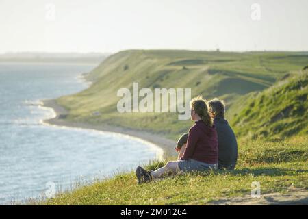 Immagine di una coppia seduta sull'erba verde sulla scogliera che guarda al mare durante il tramonto Foto Stock