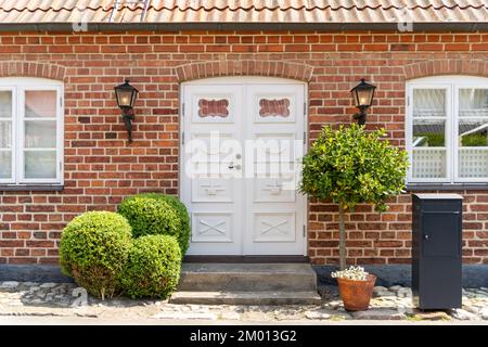 Immagine della porta d'ingresso in legno bianco vintage con lampade laterali in metallo, pareti in mattoni e verde imboscato di fronte ad essa Foto Stock