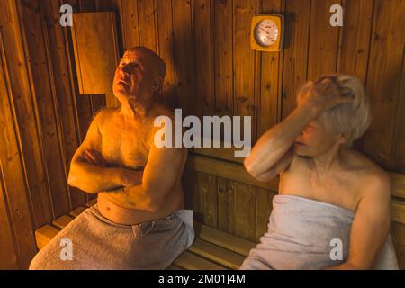 Due anziani caucasici - uomo calvo e donna a pelo corto - si siedono insieme in una sauna di legno in accappatoi di cotone grigio. CONCETTO DI BENESSERE e relax. Foto di alta qualità Foto Stock