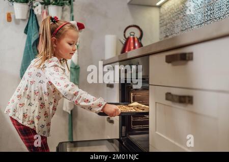 Piccola ragazza carina che fa biscotti dolci fatti in casa. Ha messo una teglia nel forno caldo. Preparazione dei biscotti di Natale. Casa dolce casa e caldo f Foto Stock