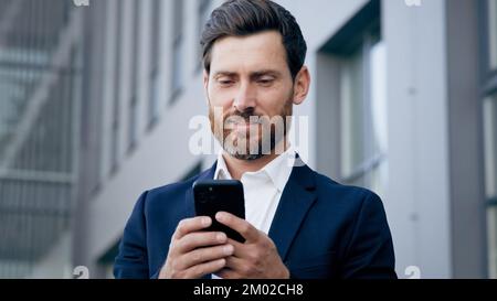 Un giovane uomo d'affari di successo che tiene uno smartphone in piedi nel centro affari della città utilizza il telefono scrive un messaggio di testo sorridendo uomo manager utilizzando il cellulare Foto Stock