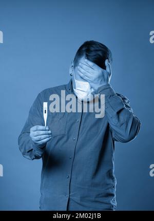 Giovane uomo in tessuto casual indossare maschera medica protettiva eccitato, stressato e spaventato dopo la lettura molto alta temperatura risultato da digitale Foto Stock
