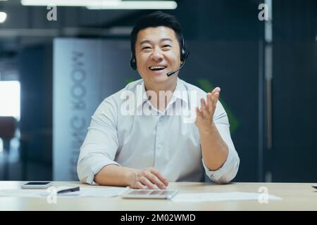 Call center. Telemarketing, assistenza clienti per la crescita delle vendite. Un giovane asiatico parla e consulta la fotocamera per il servizio clienti in cuffia. Seduto sorridendo in ufficio al tavolo. Foto Stock