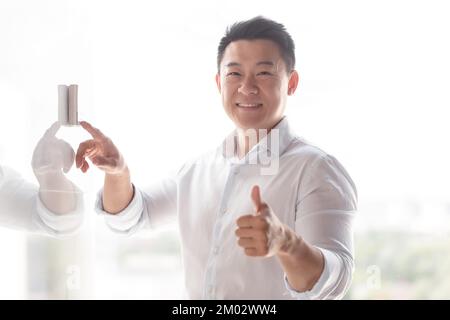 Ritratto di un uomo asiatico sorridente. Si trova accanto al muro dell'edificio, preme la chiamata in arrivo, pannello di controllo con la mano. Guarda la telecamera, mostra un super. Foto Stock