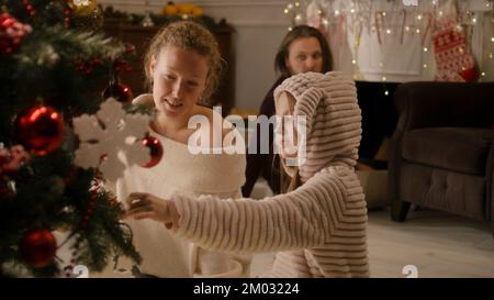 Giovane ragazza in pigiama mite scelta di palle e decorazione albero di Natale con la mamma. Papà accende il caminetto. La famiglia prepara il soggiorno prima di Natale o Capodanno. Atmosfera accogliente delle vacanze invernali. Foto Stock