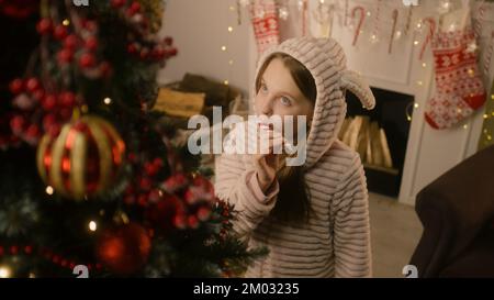 Giovane ragazza in pigiama mite viene all'albero di Natale, lecca caramelle e corregge palle e giocattoli. Il Garland risplende sul muro. Preparazione prima delle vacanze invernali. Magia del nuovo anno. Movimento lento. Foto Stock