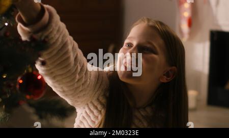 Giovane ragazza in pigiama mite siede sul pavimento, decora l'albero di Natale con palle e giocattoli, sorride e trascorre il tempo libero a casa. I bambini preparano la camera prima delle vacanze invernali e di Capodanno. Movimento lento. Foto Stock