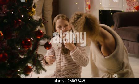 Ragazza giovane in pigiama mite decorazione albero di Natale con rami. Mamma vestire il giocattolo fiocco di neve sull'orecchio della figlia. Famiglia divertirsi e preparare il soggiorno prima delle vacanze di Natale. Movimento lento Foto Stock