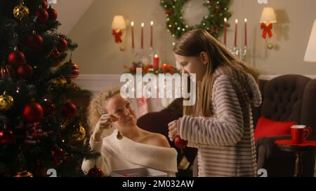 Giovane ragazza in pigiama mite scelta di palle e decorazione albero di Natale con la mamma. Papà accende il caminetto. Famiglia preparare il soggiorno prima di natale o di nuovo anno. Atmosfera accogliente delle vacanze invernali Foto Stock