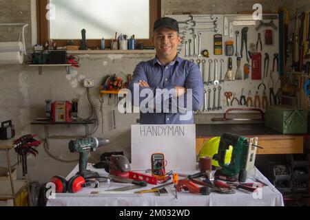 Immagine di un uomo sorridente nella sua officina con una panca piena di attrezzi di lavoro e la parola handyman. Uomo capace di riparare e fissare tutto nel h Foto Stock