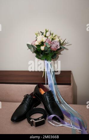 Accessori per lo sposo. Scarpe da uomo in pelle nera, bouquet di sposa, cintura in pelle nera su sfondo marrone. Foto Stock