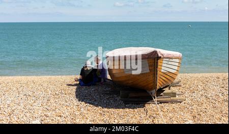 Dover, Kent, regno unito, 25, agosto 2022 uomo e donna seduti accanto ad una vecchia barca da pesca in legno su una spiaggia di ciottoli che si rilassa all'ombra Foto Stock