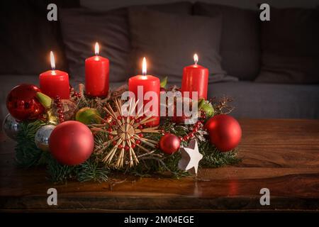 Corona dell'Avvento con quattro candele rosse brucianti e decorazione di Natale su un tavolo di legno di fronte al divano, arredamento festivo per la quarta Domenica Foto Stock