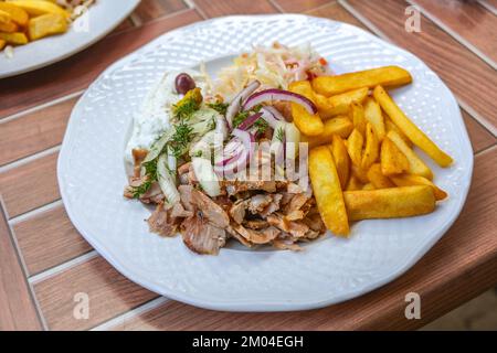 Gyros, piatto greco a fette di carne arrosto su uno spiedo, servito con patatine fritte, coleslaw, tzatziki e cipolle su un piatto bianco, all'aperto in legno Foto Stock