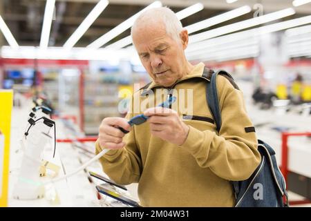 uomo anziano in grigio pensionato banco di esame con gadget elettronici e smart watch in showroom del negozio di merci digitali Foto Stock