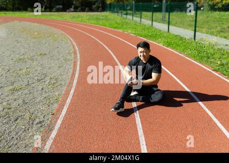 Lesioni sportive. Un giovane asiatico è seduto allo stadio su un tapis roulant, tenendo la gamba e il ginocchio. Lui si torce nel dolore. Ha bisogno di assistenza medica. Foto Stock