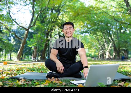 Formazione online. Bell'uomo asiatico, allenatore sportivo siede su un tappetino del parco con un computer portatile e conduce l'allenamento a distanza con una videocamera. Guarda la telecamera, sorride. Foto Stock