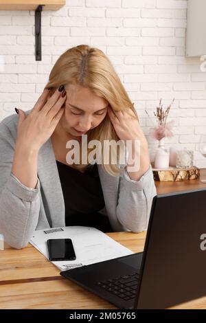 donna d'affari di mezza età, stanca e stressata, che lavora in un ufficio domestico, seduto alla scrivania accanto a un computer portatile e a documenti, pensando ai problemi online Foto Stock