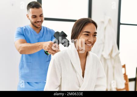 Latino uomo e donna che indossano la fisioterapia uniforme massaggiante collo con pistola a percussione al centro di bellezza Foto Stock