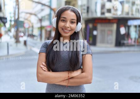 Giovane donna araba che ascolta musica in piedi con le braccia incrociate gesto per strada Foto Stock