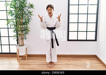 Giovane ragazza ispanica con kimono karate e cintura nera che celebra pazzo e pazzo per il successo con braccia sollevate e gli occhi chiusi urlando eccitato. vinci Foto Stock