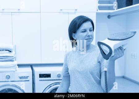 Donna stira camicia bianca a bordo in lavanderia con lavatrice sullo sfondo Foto Stock