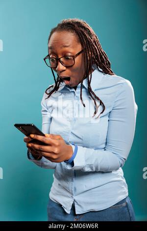 Scioccata donna afroamericana con smartphone touchscreen stupito dai messaggi ricevuti. Sorpreso eccitato giovane persona adulta con telefono moderno in shock dopo la lettura di notizie. Foto studio Foto Stock