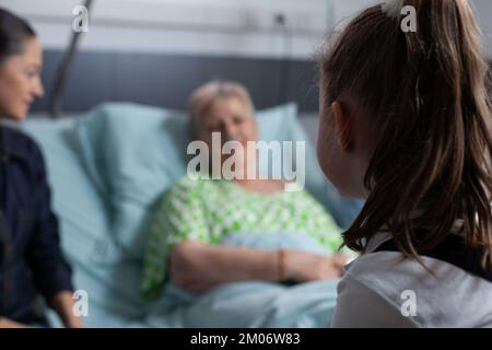 Donna anziana fuori fuoco, sdraiata nel letto di ospedale che chiacchiera con bambina non identificata. Famiglia in visita ricoverando anziana paziente geriatrica in sala clinica medica. Foto Stock