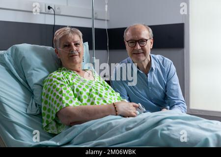 Uomo anziano che accompagna il compagno di letto in una stanza d'ospedale. Paziente femmina che riceve visita da marito in clinica medica stanza di cura. Ritratto di vecchia coppia sorridente in sanatorio. Foto Stock