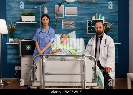 Ritratto di donna anziana sdraiata nel letto di casa di cura con un team medico che la assiste nella stanza. Medico e infermiere specializzato in assistenza agli anziani, assistenza sanitaria. Foto Stock