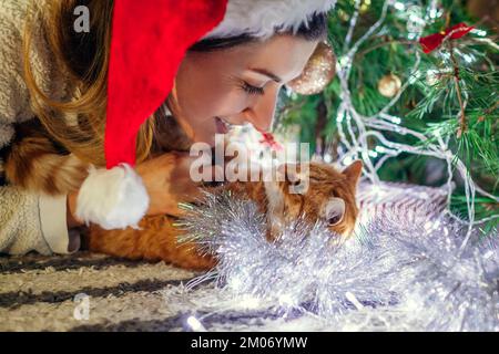 Festa di Natale con gatto. Donna che gioca e abbraccia l'animale domestico nel cappello di Babbo Natale dall'albero di Capodanno a casa. Animale coperto di tinsel e luci Foto Stock