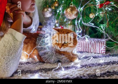 Festa di Natale con gatto. Donna ammirando l'arredamento da albero di Capodanno a casa coperto di tinsel e luci. Solo festeggiamenti Foto Stock