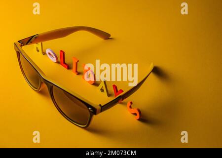 L'iscrizione Vacanze da lettere di legno luminoso multicolore giace su uno sfondo giallo brillante con occhiali da sole. Vista dall'alto. Spazio di copia. vacanza Foto Stock