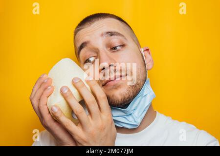 Un uomo in una camicia blu gli avvolse il volto in carta igienica, cercando di proteggersi dal coronavirus. In studio su sfondo giallo. Protezione Foto Stock